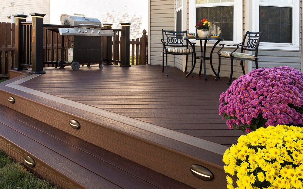 Custom-designed outdoor deck built by Zenith Builders featuring rich walnut-toned planks with a smooth finish, paired with contrasting deep blue railing and balusters. Elegant stairs descend to a verdant, tree-lined backyard, highlighting quality craftsmanship and design