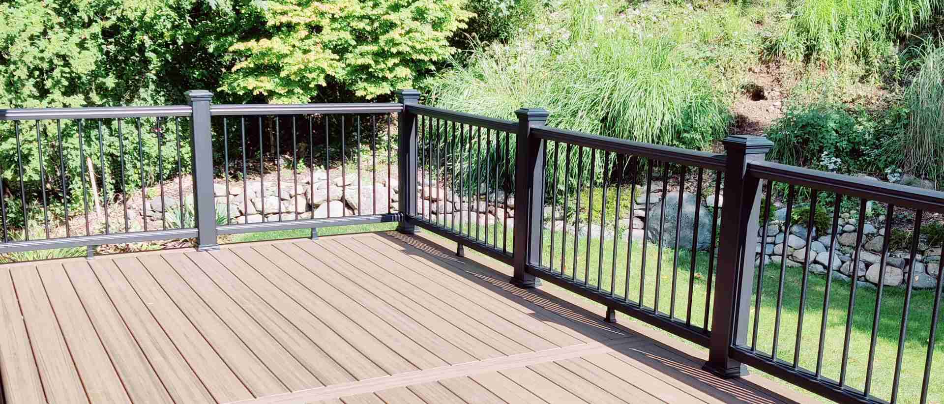 A full deck rebuild by Zenith Builders features a spacious, multi-tier design with luxurious walnut-colored composite planks and contrasting black aluminum balusters, all set against the serene backdrop of a dense, leafy forest